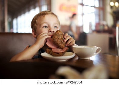 Little Boy In A Cafe During Lunch. Hungry Kid Eating Sausage From His Sandwich