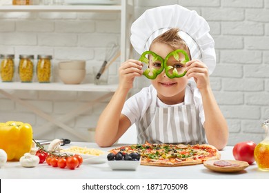 Little Boy In Chef Hat And An Apron Cooking Pizza In The Kitchen. The Child Holding Green Bell Peppers . Having Fun