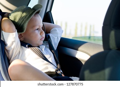 Little Boy In A Child Safety Seat Sitting Patiently In The Back Of A Car With His Hands Behind His Head Staring Out Of The Window