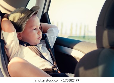 Little Boy In A Child Safety Seat Sitting Patiently In The Back Of A Car With His Hands Behind His Head Staring Out Of The Window