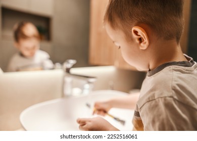 A little boy is cleaning toothbrush under the tap in a bathroom.