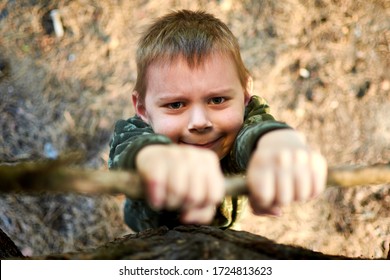 A Little Boy Climbs A Tree. Hanging With Effort On A Tree Branch.