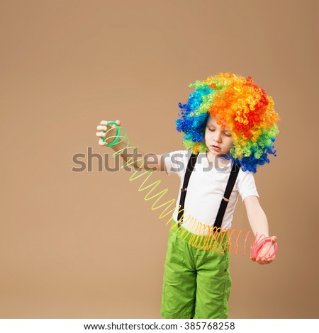 Little boy in clown wig smiling and playing with Magic Spring. Happy clown boy with large colorful wig. Birthday boy. Little clown boy with colorful hair and Slinky spring toy. 