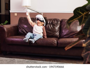 Little Boy In Comfy Clothes Using VR Set While Sitting On A Sofa. Kid Raising Hand With A Joystick.