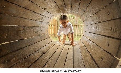 Little boy crawling through wooden tunnel or pipe on the playground. Kids playing outdoors, children having fun, summer vacation and holiday - Powered by Shutterstock