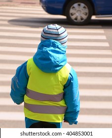 Little boy is crossing zebra crossing where any reckless driver of car pulled into. Child is wearing yellow reflective vest and jacket with reflective strips because of safety.