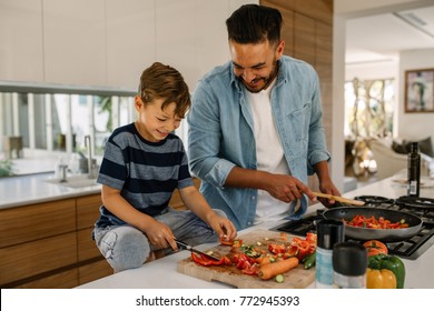 Little boy cutting vegetables while his father cooking food in kitchen. Father and son preparing food at home kitchen. Stock Photo