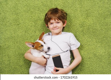 Little Boy With A Dog Listen To Music On The Phone While Lying On The Carpet. Friendship, Stay Home Concept.