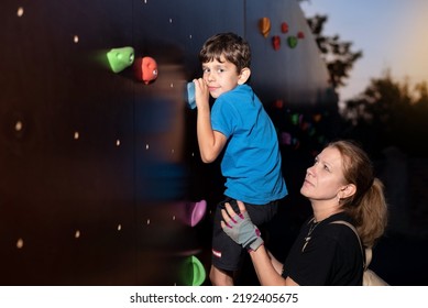 Little Boy Doing Sports With Mom In The Evening, Boy Climbs The Rock Climbing Wall, Mom Holds Him So He Doesn't Fall, Outdoor Sports. The Boy Dreams Of Being A Rock Climber