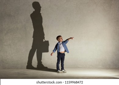 The little boy dreaming about businessman or diplomat profession. Childhood and dream concept. Conceptual image with boy and shadow of man in suit on the studio wall