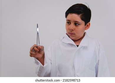 Little Boy Dressed As A Doctor On A White Background With A Medical Tool In His Hand. Boy Dressed In Doctor's Gown Holding A Medical Instrument Isolated On A White Background.