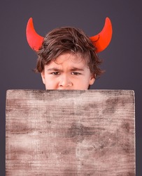 Little Boy Dressed Like A Devil Holding A Wooden Board Over Grey Background