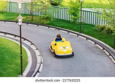 A Little Boy Is Driving A Miniature Yellow Car On The Road In The Children's Town. Racetrack For Kids In The Amusement Park