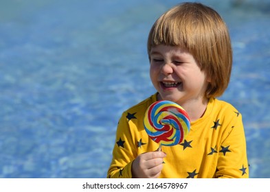 A Little Boy Eats A Big Colorful Candy Sitting By The Pool. Concept: Bright Summer, Family Vacation, Travel With Children, Resort, Water Park. Space For Text 