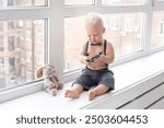 Little boy eats fruit puree from a doypack dressed in a suit with trousers on pull-ups and a shirt near blue wall at home