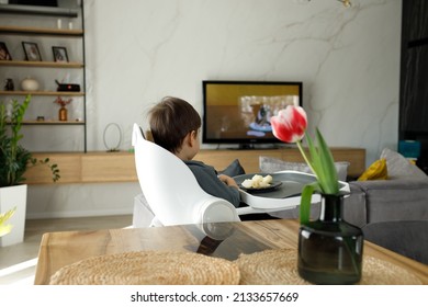 Little boy in feeding chair eating apples and bananas for breakfast in front of the cartoons on tv in a modern living room  - Shutterstock ID 2133657669