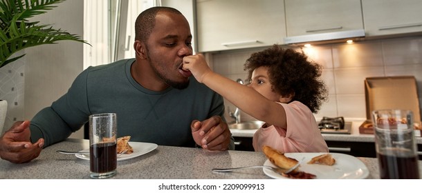 Little Boy Feeding Pizza Slice His Adult Father During Having Lunch Or Dinner At Table At Home Kitchen. Unhealthy Eating. Young Black Family Lifestyle And Relationship. Fatherhood And Parenting