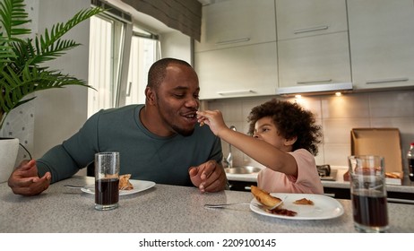 Little Boy Feeding Pizza Slice His Adult Father During Having Lunch Or Breakfast At Table At Home Kitchen. Unhealthy Eating. Young Black Family Lifestyle And Relationship. Fatherhood And Parenting