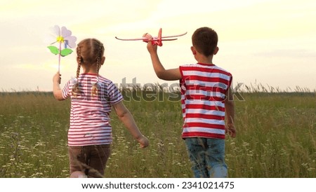 Little boy and girl with favorite toy plane and windmill flower walk on green meadow backside view. Happy little boy with girl hold toys in sunset field. Boy and girl siblings vacation in rural nature