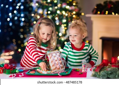 Little Boy And Girl Making Christmas Gingerbread House At Fireplace In Decorated Living Room. Kids Playing With Ginger Bread Under Christmas Tree. Baking And Cooking With Children For Xmas At Home.