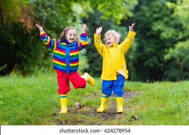 Little boy and girl play in rainy summer park. Children with colorful rainbow jacket and waterproof boots jump in puddle and mud in the rain. Kids walk in autumn shower. Outdoor fun by any weather.