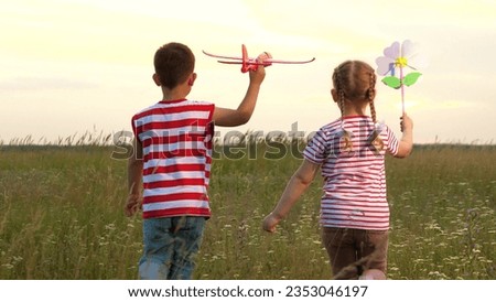 Little boy and girl play with toy plane and windmill walking on large meadow backside view. Healthy boy with girl with toys at sunset farmland. Boy and girl siblings have fun with toys in wild nature