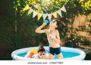 Little boy and girl, sibling, playing in inflatable pool outdoors swimming in inflatable circles on a sunny summer day. Boy blowing soap bubbles. Kids are fun and happy. Healthy lifestyle concept. - Powered by Shutterstock