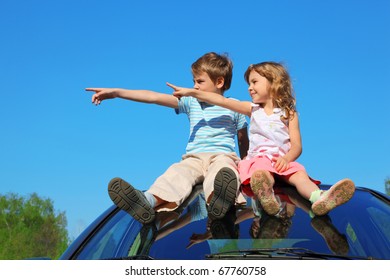 Little Boy And Girl Sitting On Car Roof On Blue Sky, Looking To Left Side And Showing By Finger