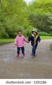 A Little Boy And Girl Wearing Wellington Boots Holding Hands And Running Through A Country Park