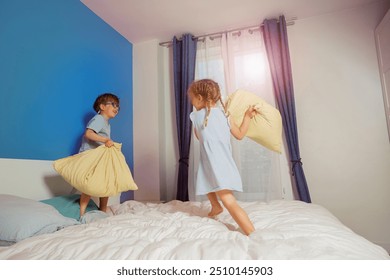 A little boy in glasses and a girl with braided hair happily engage in a pillow fight on a cozy bed - Powered by Shutterstock