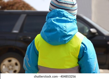 Little Boy Is Going To Cross The Road Where Green Car Is Driving. Child Is Wearing Yellow Reflective Vest And Jacket With Reflective Strips Because Of Safety.