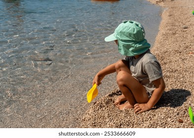 Little boy in a green cap playing with small pebbles on the seashore using a yellow spatula on a sunny day. Concept of childhood, outdoor play, and summer vacations by the sea. High quality photo - Powered by Shutterstock