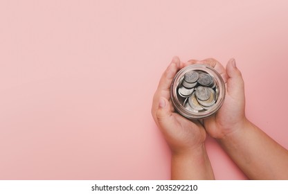 Little Boy Hand Holding Money Coin In Glass Jar On Pink Background, Donation, Saving, Fundraising Charity, Family Finance Plan Concept, Superannuation, Financial Crisis Concept, Top View.