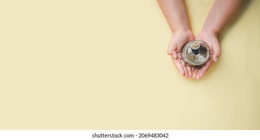 Little boy hand holding money coin in glass jar on yellow background, donation, saving, fundraising charity, family finance plan concept, superannuation, financial crisis concept, Top view.