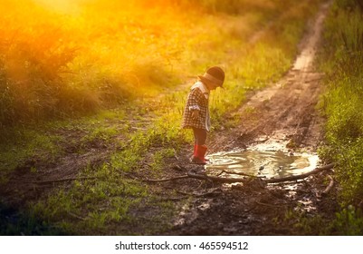 Little boy in hat and rubber shoes playing in puddle in summer forest.
