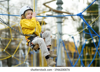 Little Boy Having Fun In Adventure Park For Children Amoung Ropes, Stairs, Bridges. Outdoor Climbing Adventure Playground In Public Park. Child Slides Down The Rope With The Help Of A Bungee.