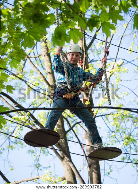Little boy in the helmet and a safety system\
stands on the suspension bridge and keeps the ropes against the sky\
and the trees with\
leaves