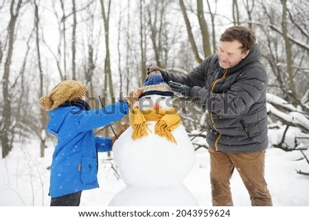 Little boy with his father building snowman in snowy park. Dad and son tied a scarf for snowman. Active outdoors leisure with family with children in winter. Kid during stroll in a snowy winter park