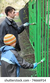Little boy and his father stand back and dye fence by green color on community work day, boy in focus