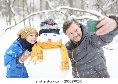 Little Boy And His Father Taking Selfie On Background Of Snowman In Snowy Park. Active Outdoors Leisure With Children In Winter. Family During Stroll In A Snowy Winter Park