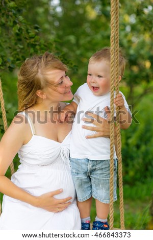 A little boy with his pregnant mother sitting on a swing. Pregnant mom with a boy resting in a meadow. Mother and son outdoors in summer