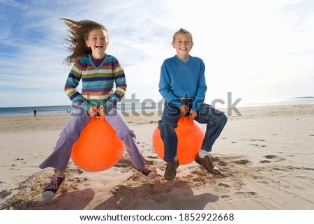 Little boy and his sister sitting on inflatable hoppers and bouncing on the beach on a bright, sunny day