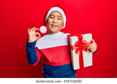 Little Boy Hispanic Kid Wearing Christmas Hat And Holding A Gift Doing Ok Sign With Fingers, Smiling Friendly Gesturing Excellent Symbol 