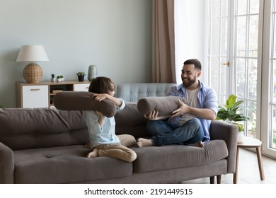 Little Boy Hold Cushion Play Fight With Young Cheery Father Sit On Sofa At Home, Happy Family Enjoy Active Pastime, Spend Leisure Together At Modern Fashionable Home. Games, Fun, Fatherhood Concept