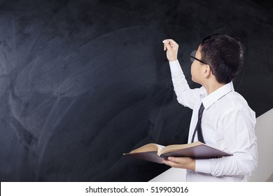 Little boy holding a book and writing on the empty chalkboard with a chalk, shot in the class