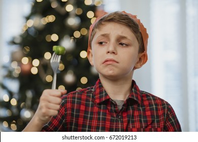 Little Boy Is Holding A Brussel Sprout On His Fork At Christmas Time. He Is Wearing A Party Hat And Is Grimacing At The Vegetable. 