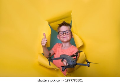 Little Boy Holding Drone And Breaking Through Yellow Paper Wall. Kid With Radio Remote Control For Drone. Smart School Boy With Thumb Up. Nerd Child In Glasses.