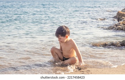 Little Boy Kid Having Fun In Sea Ocean  Water Lying On Sands Beach Splashing Hitting Waves With Hand Putting Sand On Legs Belly Tummy Playing Smiling.happy Child Enjoy Vacation Sunny Summer Day