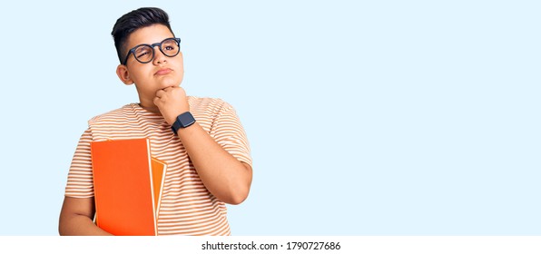 Little Boy Kid Holding Book Wearing Glasses With Hand On Chin Thinking About Question, Pensive Expression. Smiling With Thoughtful Face. Doubt Concept. 