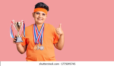 Little Boy Kid Holding Champion Trophy And Wearing Medals Smiling Happy And Positive, Thumb Up Doing Excellent And Approval Sign 